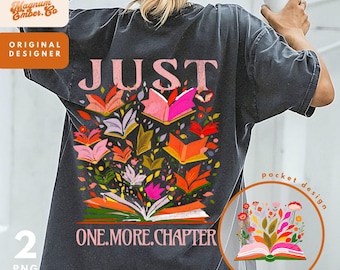May include: A black t-shirt with a graphic design of a book with flowers and the text "JUST ONE.MORE.CHAPTER". The pocket design features a similar book with flowers.