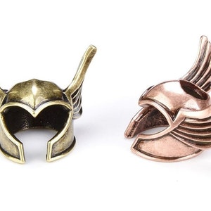 May include: Two miniature winged helmets, one in gold and one in copper, are shown against a white background. The helmets are detailed with wings and a raised design.