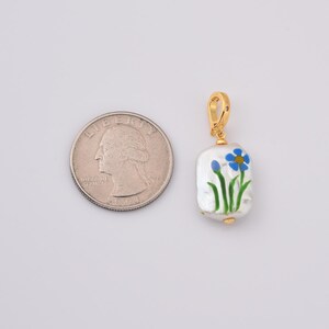 May include: A gold-colored pendant with a rectangular, white pearl base. The pearl is hand-painted with a blue flower design and green stems. The pendant is shown next to a U.S. quarter for size comparison.