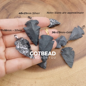May include: Five arrowhead pendants in various sizes, with silver and gold accents. The pendants are made of black obsidian and are displayed on a wooden surface. The text "GOTBEAD BEAD FOR YOU" is visible in the image.