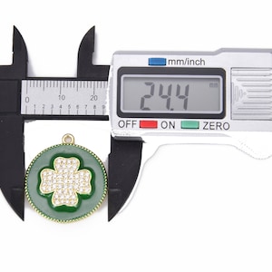 May include: A close-up of a green and gold charm with a four-leaf clover design. The charm is 2.44 cm in diameter and is covered in clear rhinestones. The charm is being measured with a digital caliper.