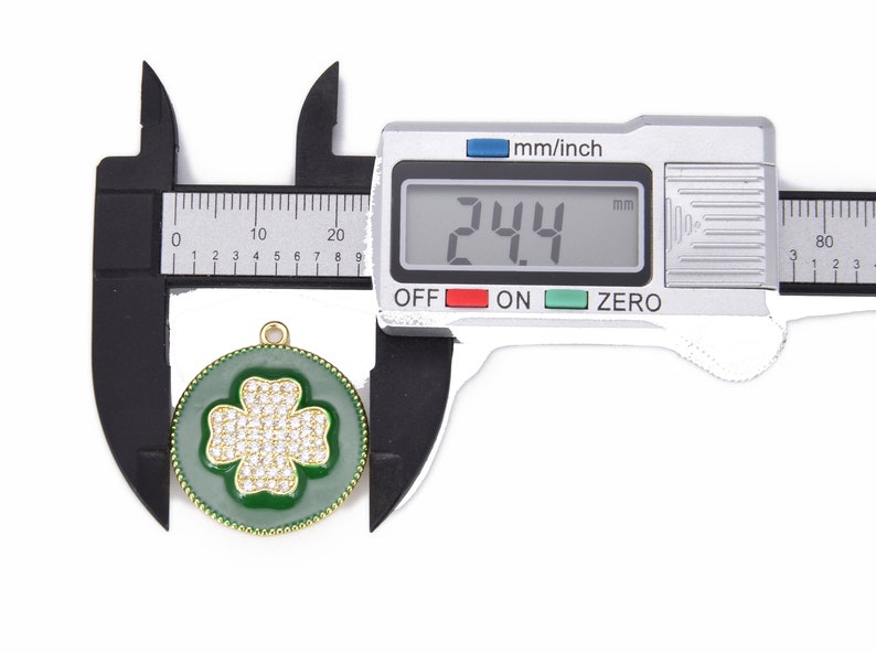 May include: A close-up of a green and gold charm with a four-leaf clover design. The charm is 2.44 cm in diameter and is covered in clear rhinestones. The charm is being measured with a digital caliper.