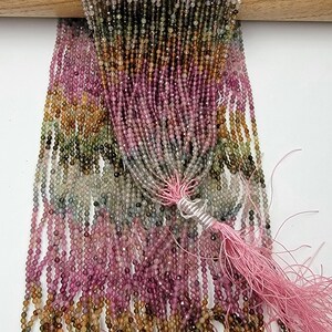 May include: A collection of multi-colored tourmaline gemstone beads arranged in long, thin strands. The beads feature a faceted finish and display a gradient of pink, green, yellow, and brown hues. A light pink thread tassel is tied at the bottom.