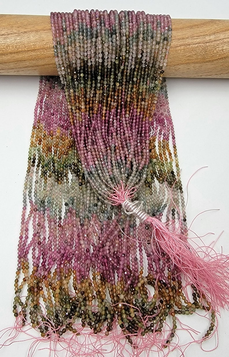 May include: A collection of multi-colored tourmaline gemstone beads arranged in long, thin strands. The beads feature a faceted finish and display a gradient of pink, green, yellow, and brown hues. A light pink thread tassel is tied at the bottom.