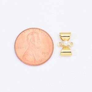 May include: A gold-toned metal clasp with a small white pearl in the centre. The clasp is shaped like a bow and has two loops on either side. The clasp is shown next to a US penny for size comparison.