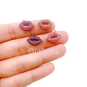 May include: Four charm pendants in the shape of lips, each with a different colored metal setting. The lips are covered in small red rhinestones. The charms are displayed on a hand.