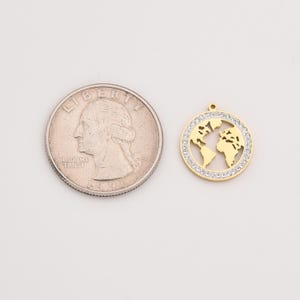 May include: A gold-colored pendant in the shape of a world map, surrounded by a ring of small, clear crystals. The pendant is next to a U.S. quarter coin for size comparison. The pendant has a small loop at the top for a chain.