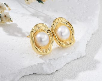 May include: A pair of gold-colored stud earrings, each featuring a large, lustrous white pearl set within a textured, organic-shaped frame. The earrings are presented on a white surface, with delicate floral accents in the background.