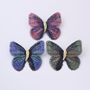 May include: Three embroidered butterfly brooches in shades of purple, blue, and green. Each butterfly has a gold-colored center and detailed wing patterns. The brooches are set against a white background.