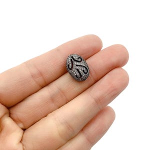 May include: An oval-shaped bead with a dark gray base, encrusted with small, sparkling stones. The bead features a black, swirling design. This jewelry component is ideal for crafting necklaces, bracelets, and earrings.