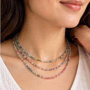 May include: A triple-strand necklace featuring small, faceted, multicolored gemstone beads in shades of pink, green, and blue. The delicate strands are arranged in a layered style, resting against the neck with a subtle, polished finish.