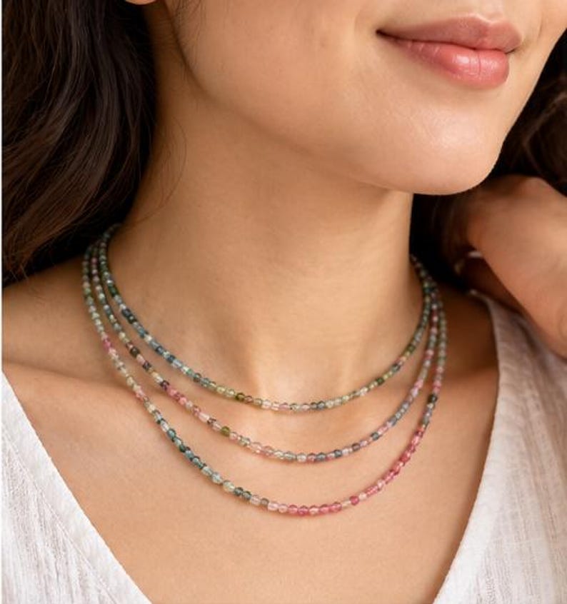 May include: A triple-strand necklace featuring small, faceted, multicolored gemstone beads in shades of pink, green, and blue. The delicate strands are arranged in a layered style, resting against the neck with a subtle, polished finish.