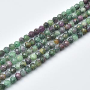 May include: A strand of faceted round beads featuring a mix of natural green and deep purple tones. Each bead displays a polished, multi-faceted surface that reflects light, creating a textured appearance across the entire length of the string.