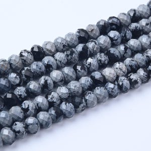 May include: Strands of faceted snowflake obsidian beads. The beads are a mix of black, gray, and white, with a speckled pattern. The beads are strung together, ready for jewelry making.