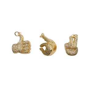 May include: Three gold-tone charms in the shape of hands. The first charm is a thumbs up, the second charm is a peace sign, and the third charm is an OK sign. All charms are covered in clear rhinestones.