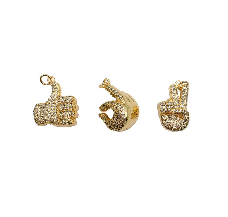 May include: Three gold-tone charms in the shape of hands. The first charm is a thumbs up, the second charm is a peace sign, and the third charm is an OK sign. All charms are covered in clear rhinestones.