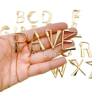May include: Gold-toned metal alphabet letter charms. The letters P, A, V, and E are displayed in a hand. Other letters are scattered around the hand. The letters are all uppercase.