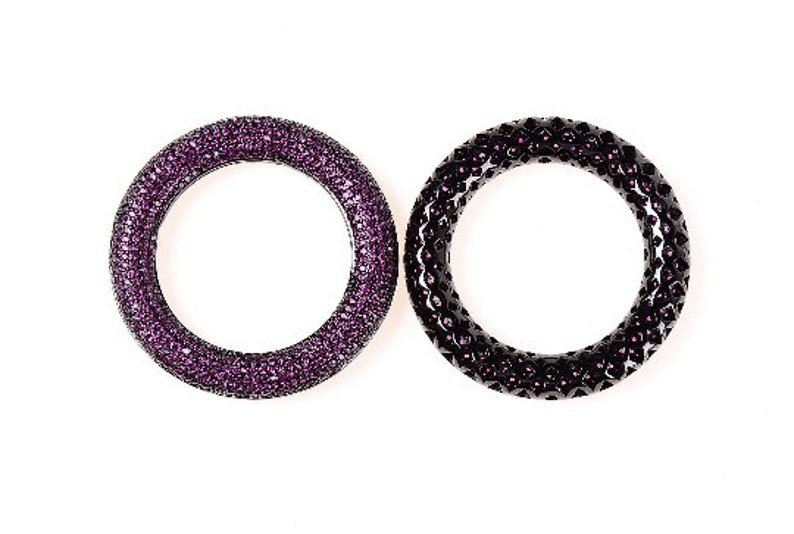 May include: Two circular bracelets. One is a deep purple, covered in small, sparkling crystals. The other is black with a textured surface and small purple crystals. Both are set against a white background.