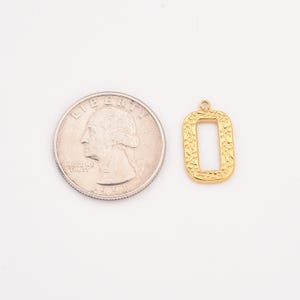 May include: A gold-colored rectangular pendant with an open center, next to a U.S. quarter. The pendant has a textured design around the edges and a small loop at the top for attaching to a chain or other jewelry component.