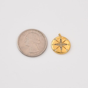 May include: A gold-colored compass pendant with a quarter coin for size comparison. The pendant features a compass rose design with embedded clear crystals. The letters N, E, S, and W are visible on the pendant. The pendant is round and has a small loop at the top.
