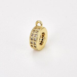 May include: Gold-toned metal charm with a round shape and a loop at the top. The charm is decorated with small clear stones.