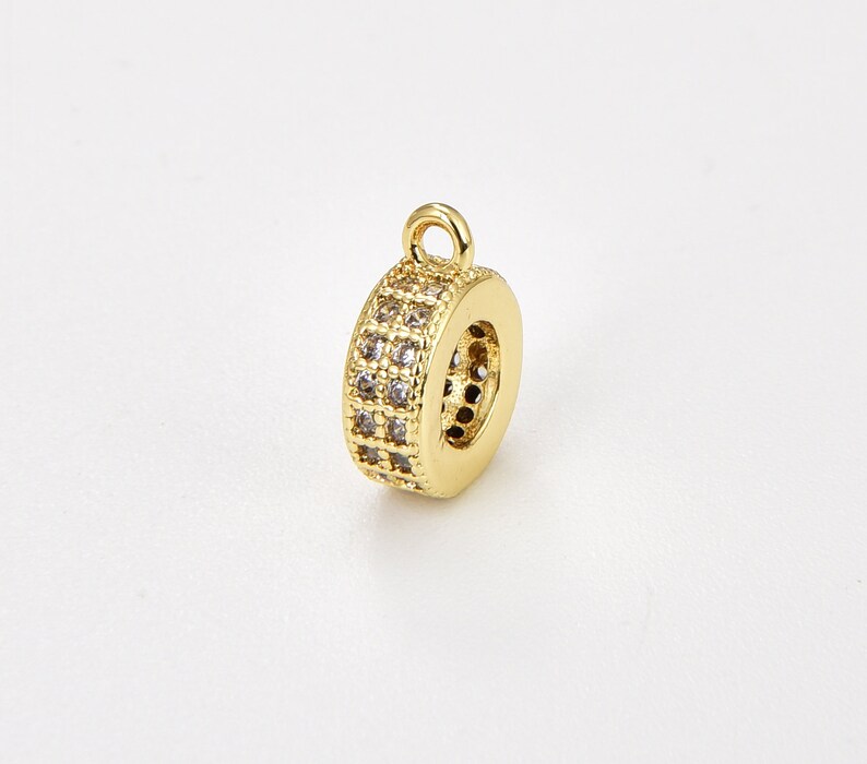 May include: Gold-toned metal charm with a round shape and a loop at the top. The charm is decorated with small clear stones.