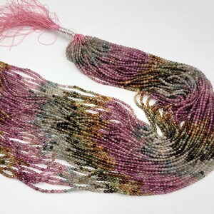 May include: A long strand of small, faceted, multi-colored tourmaline beads in shades of pink, green, brown, and grey. The beads are strung together and finished with a light pink thread tassel at one end, secured by a silver-colored wire wrap.