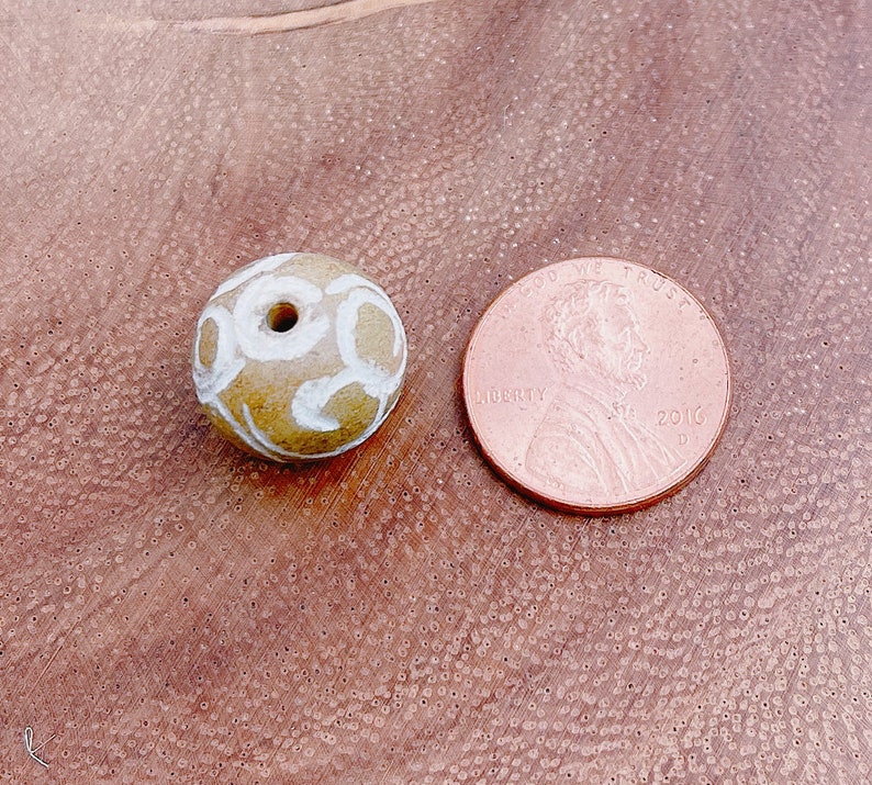 以下が含まれることがあります： 白く渦巻く模様が施された丸い茶色の陶器のビーズ。ビーズの中央には紐を通すための穴が開いています。ビーズは日本の1円玉と同じくらいの大きさです。