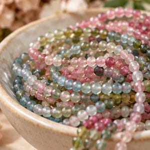 May include: A collection of multi-colored gemstone bead strands in shades of pink, green, and blue, coiled inside a speckled beige ceramic bowl. The small, round, translucent beads feature a faceted surface that reflects light.