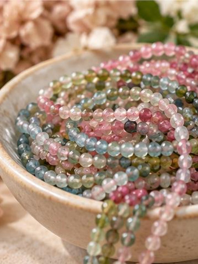 May include: A collection of multi-colored gemstone bead strands in shades of pink, green, and blue, coiled inside a speckled beige ceramic bowl. The small, round, translucent beads feature a faceted surface that reflects light.