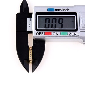 May include: A silver digital caliper measuring a small gold ring with clear stones. The caliper reads 0.09 inches.