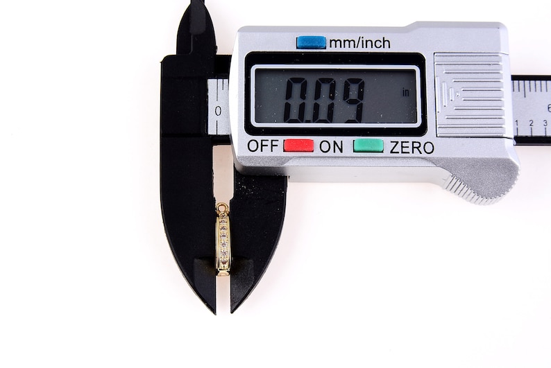 May include: A silver digital caliper measuring a small gold ring with clear stones. The caliper reads 0.09 inches.