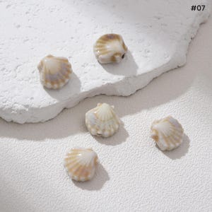 May include: Five seashell-shaped beads in shades of cream and tan are scattered on a textured white surface. Each bead has a ridged texture and a small hole for stringing. The image includes the text "#07".