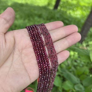 May include: A collection of strands of red garnet beads, varying in size, displayed on a hand. The beads are faceted and have a shiny, polished finish.