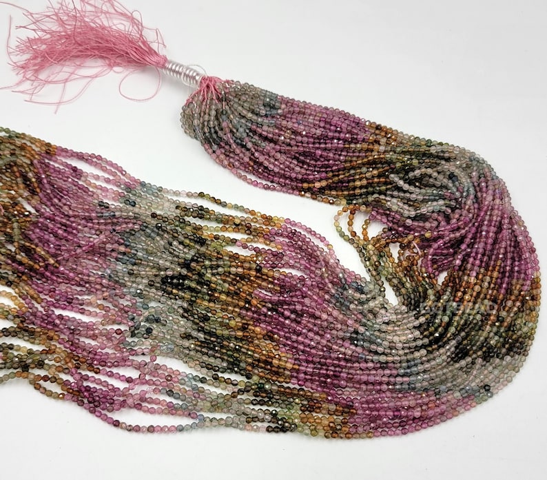 May include: A long strand of small, faceted, multicolored tourmaline beads in shades of pink, green, brown, and clear. The beads are strung on multiple threads and gathered at one end with a pink tassel tied with a silver-colored wire.