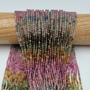 May include: A collection of numerous thin strands of small, faceted, multicolored gemstone beads. The beads feature a gradient of translucent hues including pink, green, yellow, and brown, arranged in vertical rows draped over a wooden rod.