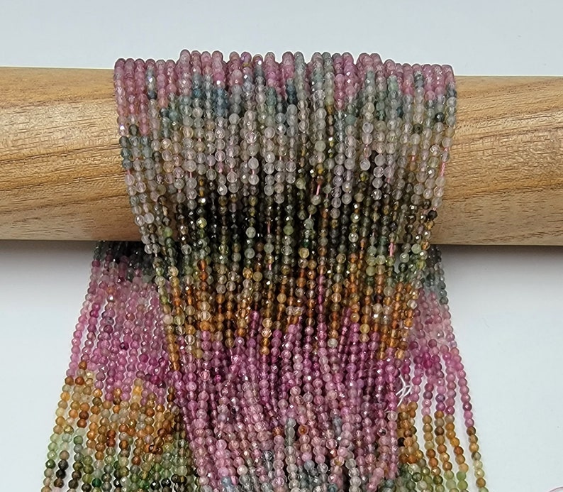 May include: A collection of numerous thin strands of small, faceted, multicolored gemstone beads. The beads feature a gradient of translucent hues including pink, green, yellow, and brown, arranged in vertical rows draped over a wooden rod.