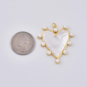 May include: A heart-shaped pendant with a mother-of-pearl center and a gold-colored frame. The frame is adorned with small, round pearls. A U.S. quarter is next to the pendant for size comparison.