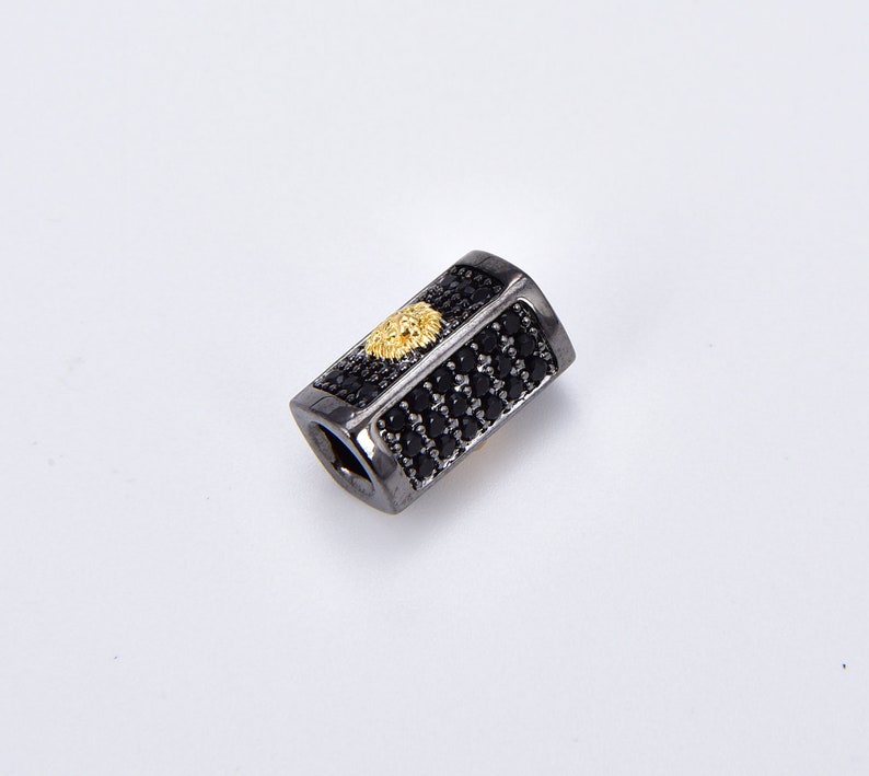 K&ouml;nnte beinhalten: Schwarze und goldene rechteckige Perle mit einem L&ouml;wenkopf-Design. Die Perle ist mit kleinen schwarzen Steinen bedeckt.