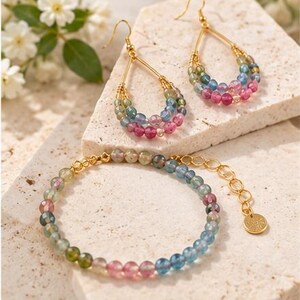 May include: A pair of gold-toned teardrop earrings and a matching beaded bracelet featuring multi-colored, faceted tourmaline gemstones in shades of pink, blue, and green. The jewelry set includes gold-toned wire accents and a small circular charm.