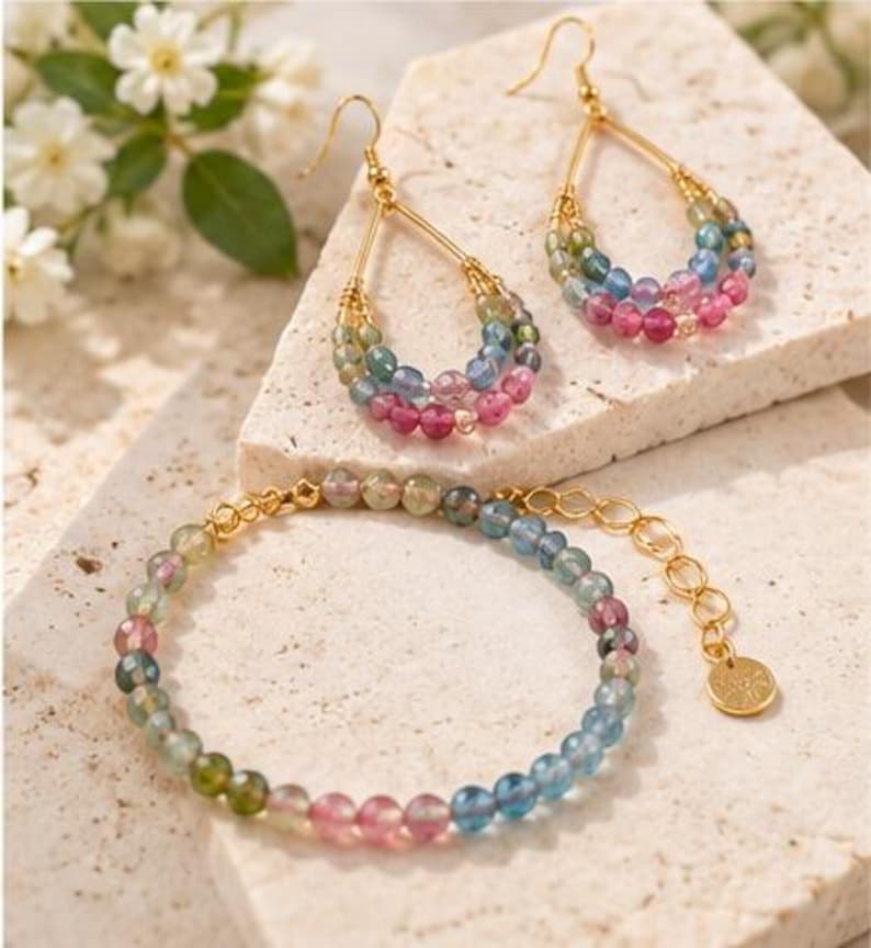 May include: A pair of gold-toned teardrop earrings and a matching beaded bracelet featuring multi-colored, faceted tourmaline gemstones in shades of pink, blue, and green. The jewelry set includes gold-toned wire accents and a small circular charm.