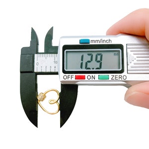 May include: A silver digital caliper measuring a gold heart charm with a small clear rhinestone. The caliper reads 1.29 mm.