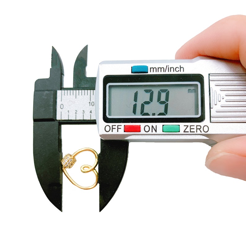 May include: A silver digital caliper measuring a gold heart charm with a small clear rhinestone. The caliper reads 1.29 mm.