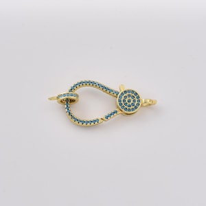 May include: A gold-toned jewelry clasp shaped like a carabiner, featuring a hinged opening mechanism. The entire frame and the circular decorative element on the side are encrusted with small, bright blue stones set in a pavé style.