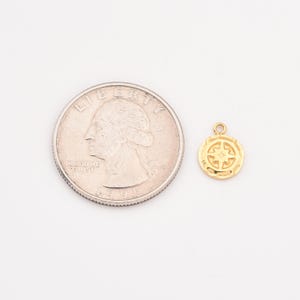May include: A small, gold-colored compass charm with a star design, next to a quarter for size comparison. The charm has a textured border and a loop for attaching to a necklace or bracelet. The quarter is silver and shows a profile of a person.