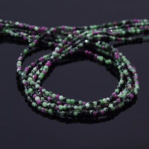 May include: A strand of green, pink, and black gemstone beads. The beads are faceted and have a round shape. The strand is coiled on a black surface.