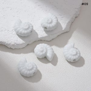 May include: Five white ceramic seashell beads with a glossy finish are arranged on a textured white surface. Each bead is shaped like a nautilus shell and has a small hole for stringing. The image includes the text "#09".