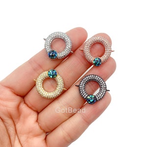 May include: Four round jewelry clasps in silver, rose gold, gold, and black. Each clasp is encrusted with small, clear stones and features a central, iridescent, round stone. The clasps are displayed against a light background.