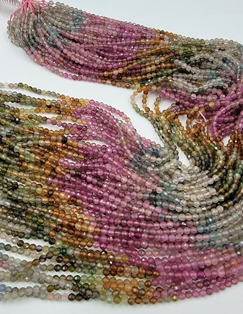 May include: A collection of multi-colored tourmaline gemstone beads arranged in multiple strands. The small, faceted, round beads display a gradient of pink, green, yellow, and blue hues, strung together on a thin, light-colored thread.