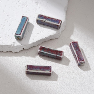 May include: Five rectangular ceramic beads with a glossy finish. The beads are primarily burgundy with teal and white accents. Each bead has a hole running through the center, suggesting they are for stringing. The beads are likely for jewelry making.